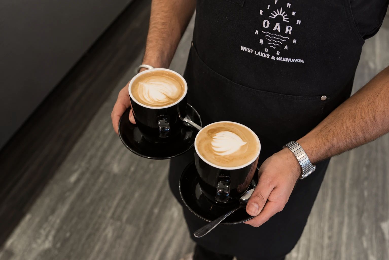 Two lattes with leaf art carried to a table by a barista in an Oar apron embroidered West Lakes and Glenunga