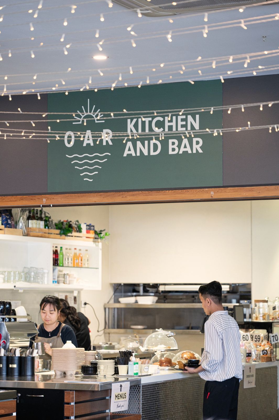 The counter at Oar Glenunga under the green Oar Kitchen and Bar sign and festoon lights, a barista at the machine and a customer ordering